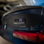 Close-up of BMW X4M Competition badge and carbon lip spoiler, highlighting M branding and carbon weave